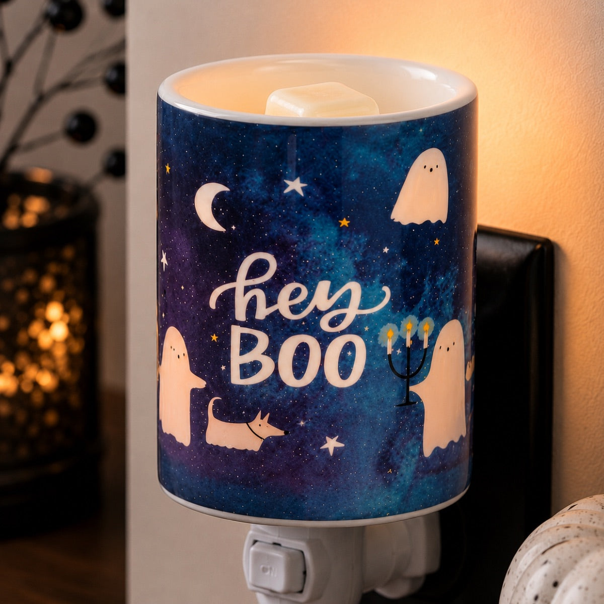 A cylindrical night light plug-in wax warmer with a blue and purple galaxy print, featuring ghost, moon, star, and 'hey BOO' illustrations. The light is turned on, casting a warm glow, and is shown plugged into a wall socket in a home setting.