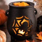 A black cauldron-shaped wax warmer with a spiderweb and crown cutout, glowing with warm orange light, filled with wax melts and set on a wooden surface with autumn decor.