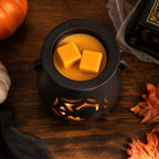 A black cauldron-shaped wax warmer with cutout details, filled with orange wax and two yellow wax cubes, styled on a dark wooden table with autumn leaves, a pumpkin, and a book.