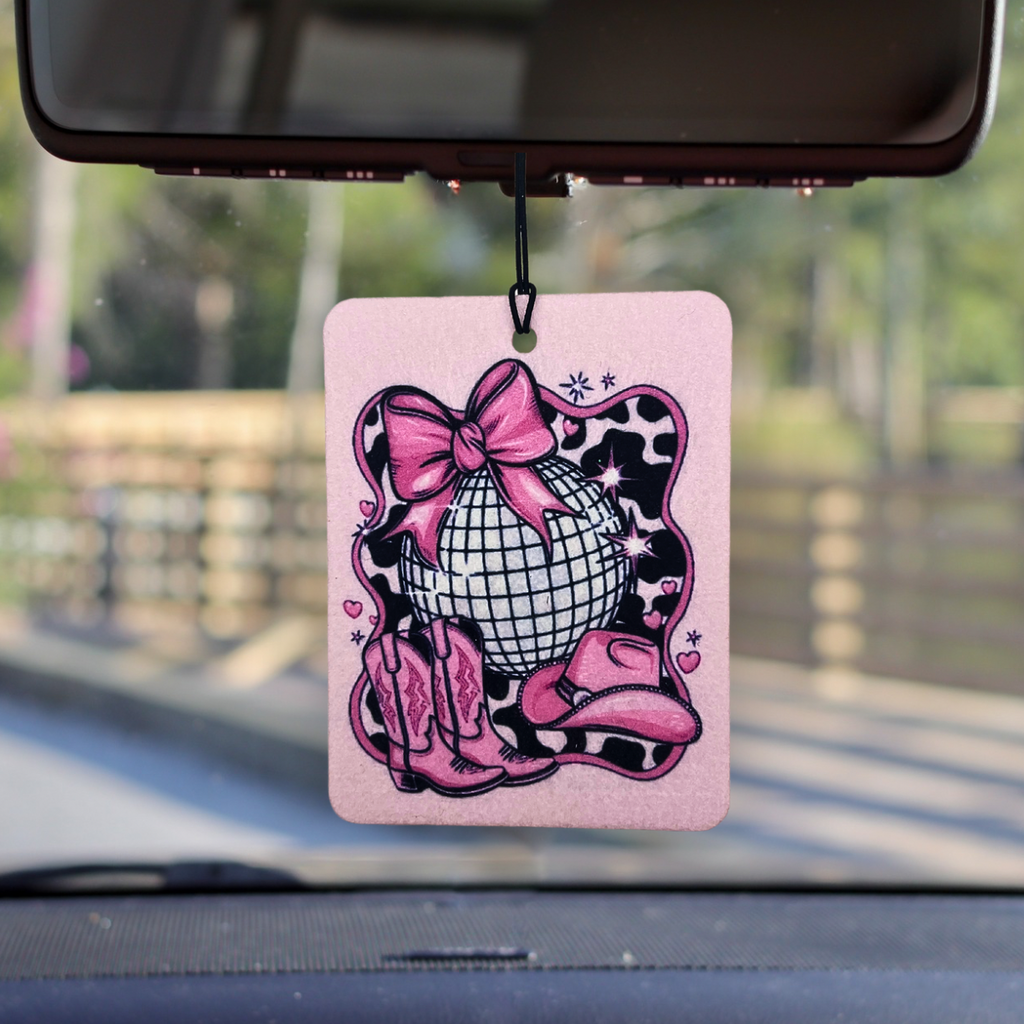 Pink car air freshener with disco ball, cowboy boots, and bow on a car dashboard.