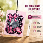Pink air freshener with a disco ball and cowboy boots design, featuring a refreshing spray on a wooden surface with a blurred floral background.