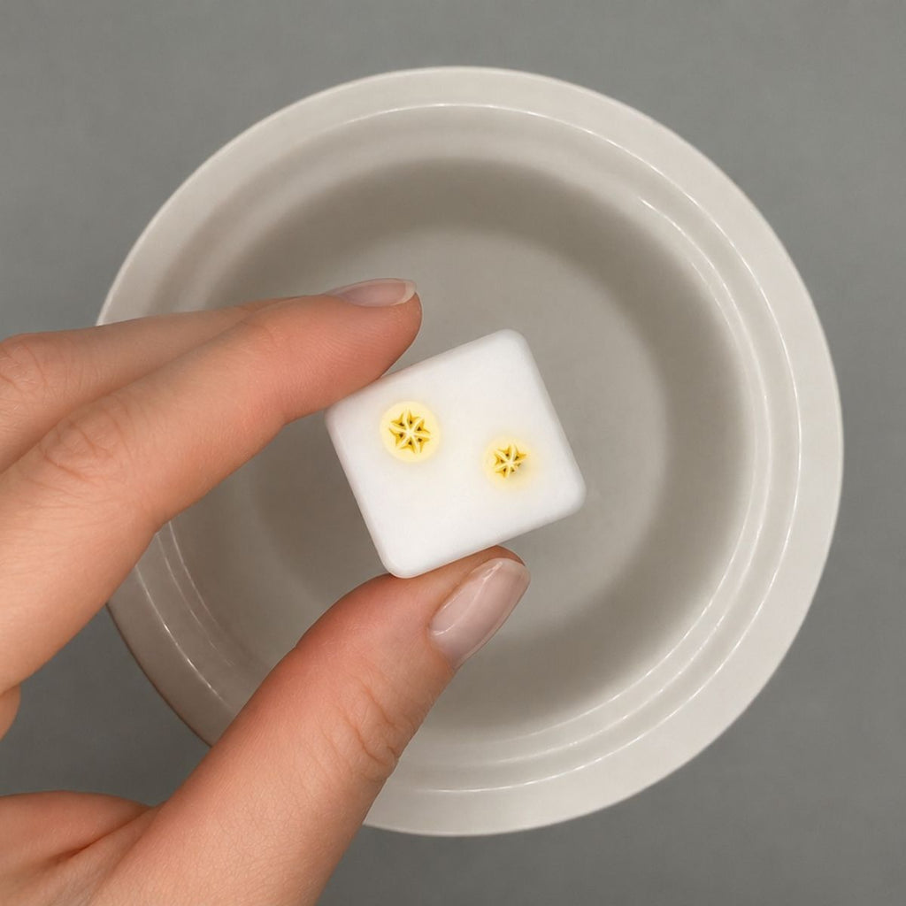 Hand holding a small white wax cube with yellow banana design on it against a gray background.