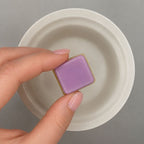 Hand holding a purple wax cube with a white wax warmer dish in the background