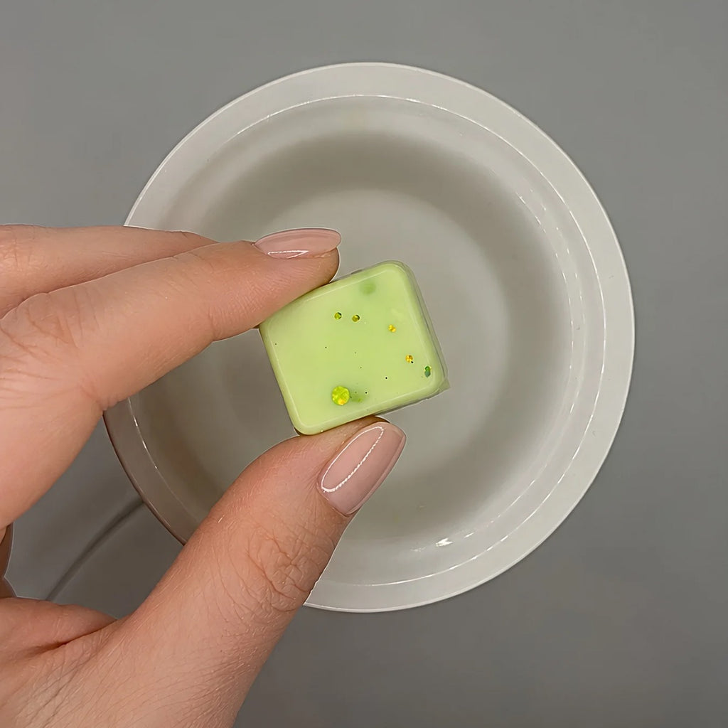 Hand holding a green wax melt cube with over a white warmer dish on a gray background