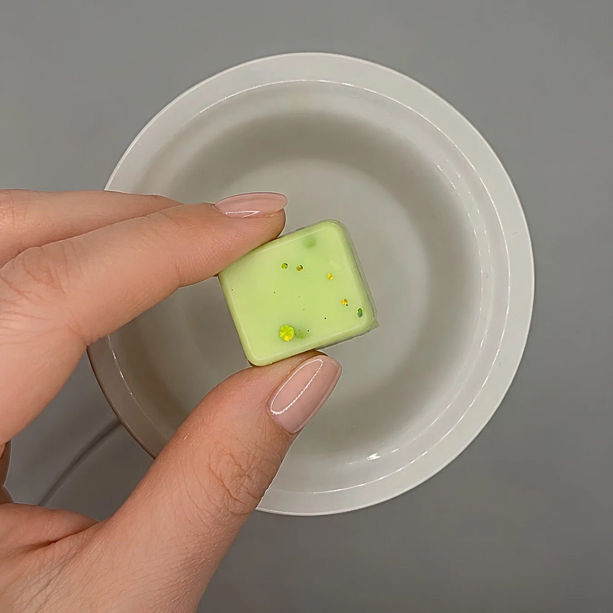 Hand holding a green wax melt cube with over a white warmer dish on a gray background