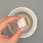 A hand holds a small square white wax melt  with orange speckles and a tan side, positioned above a small white ceramic dish on a gray background.