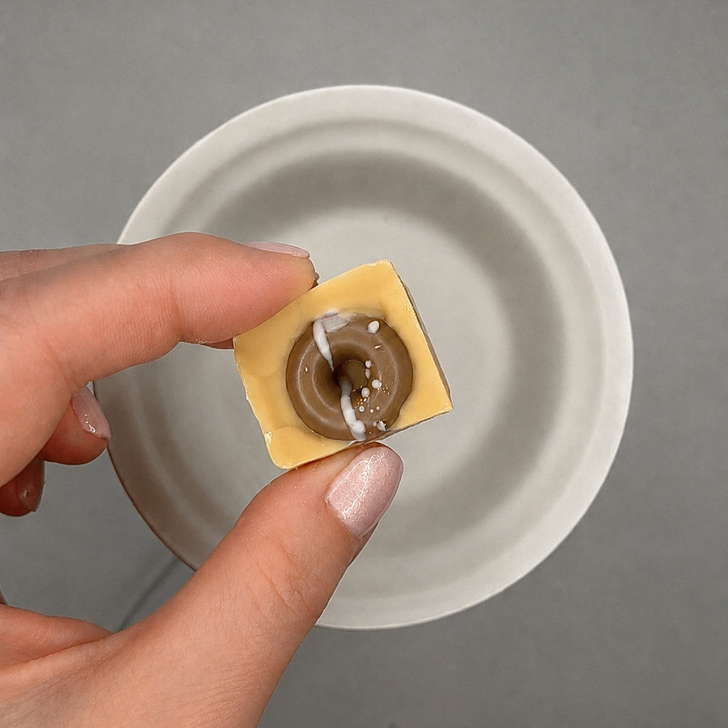 A hand holds a small square piece of a beige wax melt with a brown donut shaped center and white drizzle, positioned over a white ceramic bowl on a gray background.