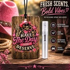 A wooden car fragrance tag with a western-themed skull and rose graphic reading 'HAVE THE DAY YOU DESERVE,' shown next to a small clear spray bottle. The scene includes a pink cowboy hat, rope, boots, and a blurred outdoor background, with text advertising 'Fresh Scents.