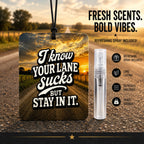 A square car air freshener hanging tag with a black cord, featuring the text 'I know YOUR LANE Sucks BUT STAY IN IT.' over a sunset road scene.