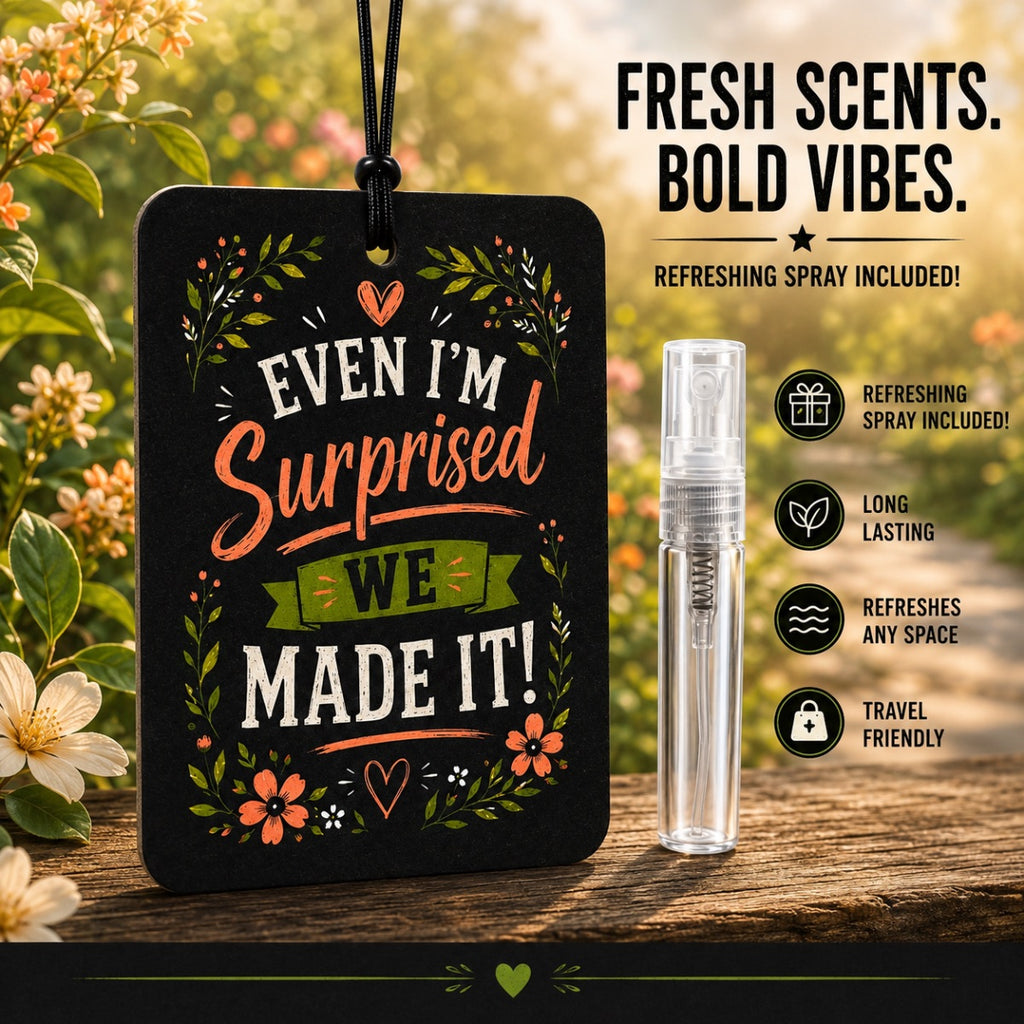 A black rectangular car air freshener tag with floral artwork and the phrase 'Even I'm Surprised We Made It!' stands on a wooden surface outdoors, next to a small clear spray bottle. Text on the right says 'Fresh Scents.
