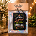 Mama Bears Scent Boutique air freshener gift set in a clear plastic bag with an iridescent holographic backing. The set includes a black rectangular air tag with the phrase 'EVEN I'M Surprised WE MADE IT!' in white, orange, and green lettering, and a small clear spray bottle.