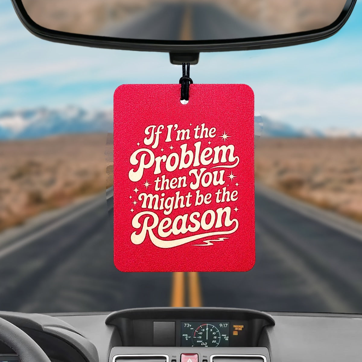 Pink car air freshener with humorous text hanging from a car's rearview mirror with a scenic road in the background.