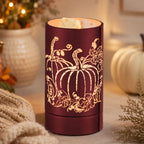 A cylindrical amber-red wax warmer candle with a laser-cut pumpkin and autumn leaf pattern, lit with warm light, set on a tabletop with cozy fall decor in the background.