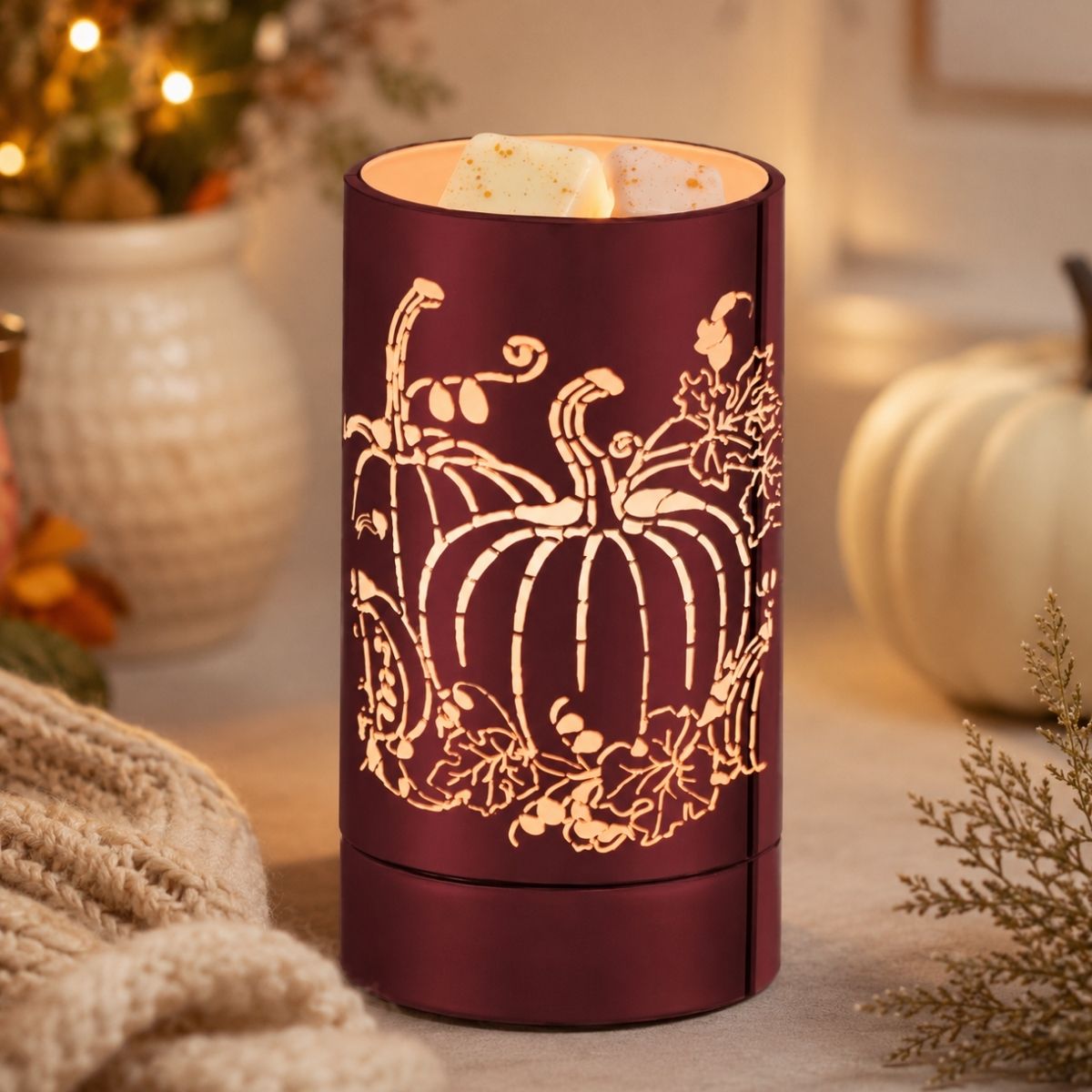 A cylindrical amber-red wax warmer candle with a laser-cut pumpkin and autumn leaf pattern, lit with warm light, set on a tabletop with cozy fall decor in the background.