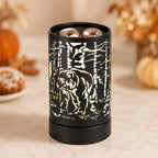 A black cylindrical wax warmer with a laser-cut forest scene featuring a bear, shown on a beige marble surface with blurred autumn pumpkins and dried flowers in the background.