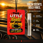 A red car air freshener spray bottle with a black hanging cord is shown on a car dashboard next to a clear travel spray vial. The background is a blurred sunset road scene.