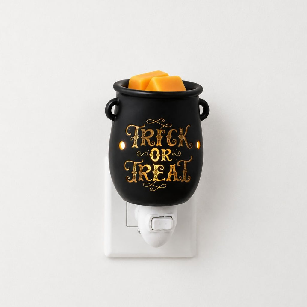 A black cauldron-shaped plug in wax warmer with gold 'Trick or Treat' lettering, filled with orange wax melts, plugged into a white wall outlet against a plain white background.