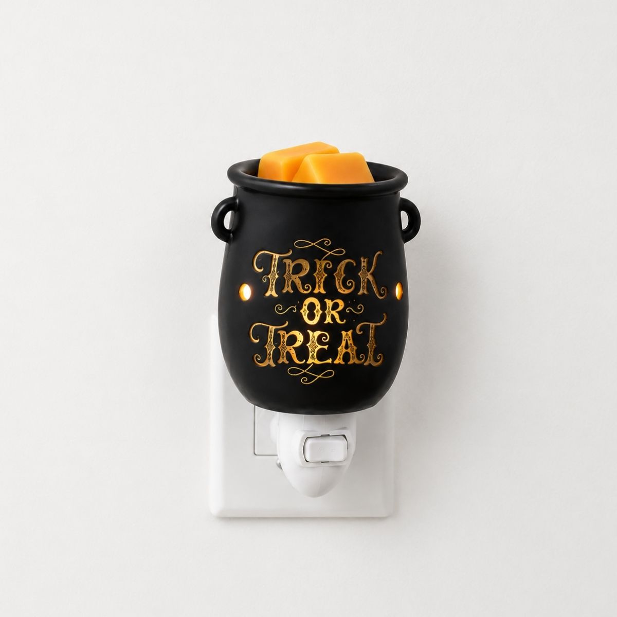 A black cauldron-shaped plug in wax warmer with gold 'Trick or Treat' lettering, filled with orange wax melts, plugged into a white wall outlet against a plain white background.