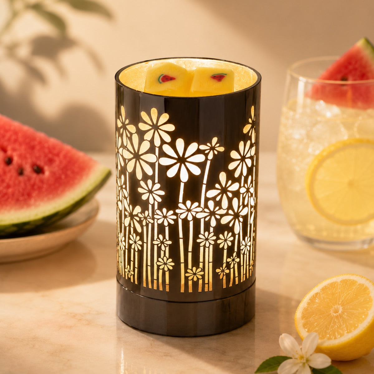 Decorative wax warmer with floral pattern on a table with fruit and a glass and two cubes of watermelon lemonade wax melts in the warmer.